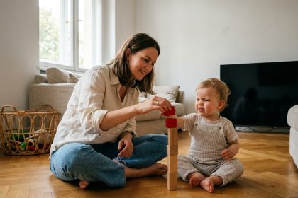 Mère et enfant en bas âge construisant une tour de cubes en bois colorés sur le parquet d'un salon, télévision éteinte en arrière-plan